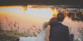 sitting woman leaning on man's shoulder facing lake during golden hour