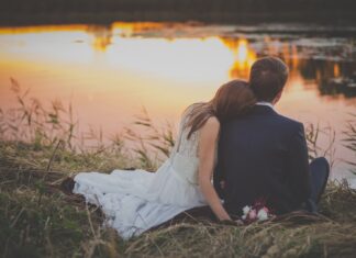 sitting woman leaning on man's shoulder facing lake during golden hour