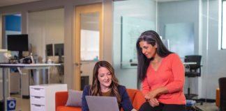 Two women on an orange couch, focused on their laptops, discussing Knowledge Management in a corporate environment.