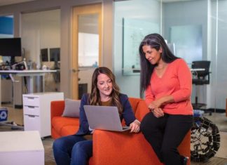 Two women on an orange couch, focused on their laptops, discussing Knowledge Management in a corporate environment.