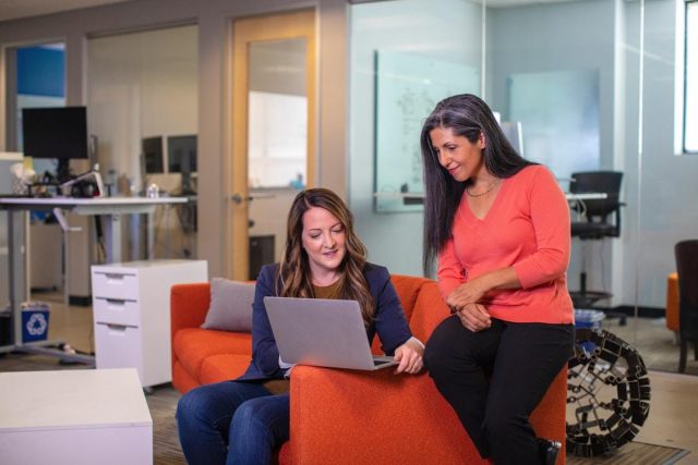 img Two women on an orange couch, focused on their laptops, discussing Knowledge Management in a corporate environment.