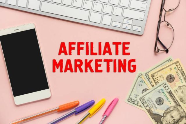 Flat lay of a smartphone, a keyboard, eyeglasses, colorful pens, and U.S. dollar bills on a pink background with the text 'Affiliate Marketing' in bold red letters. This image represents the concept of affiliate marketing, online income, and digital business opportunities.