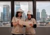 Two women in hard hats review blueprints indoors.