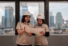 Two women in hard hats review blueprints indoors.
