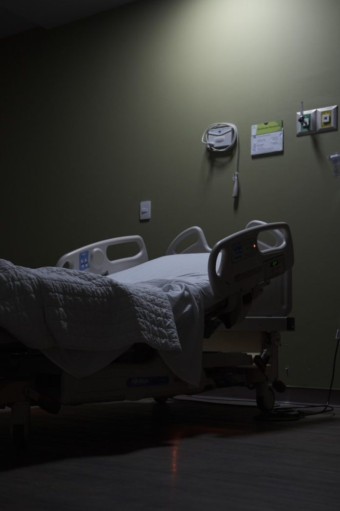 Photo by Stephan Liedtke An empty hospital bed sits in a dimly lit room.