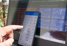 A hand pointing at a smartphone displaying currency quotes against a laptop