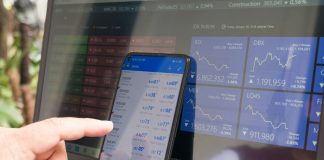 A hand pointing at a smartphone displaying currency quotes against a laptop