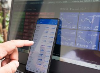 A hand pointing at a smartphone displaying currency quotes against a laptop