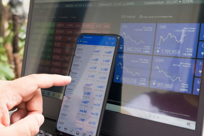 Photo by Marga Santoso A hand pointing at a smartphone displaying currency quotes against a laptop